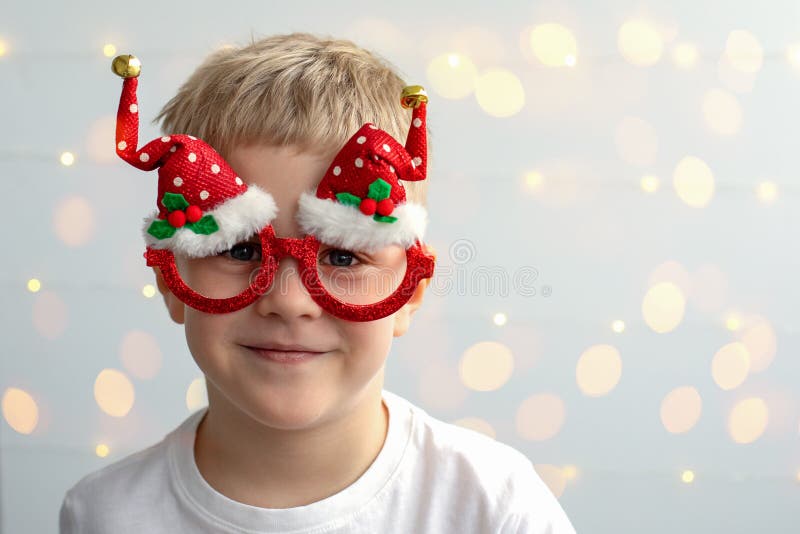 Cute smiling boy with blonde hair in festive Christmas glasses on a light background with bokeh