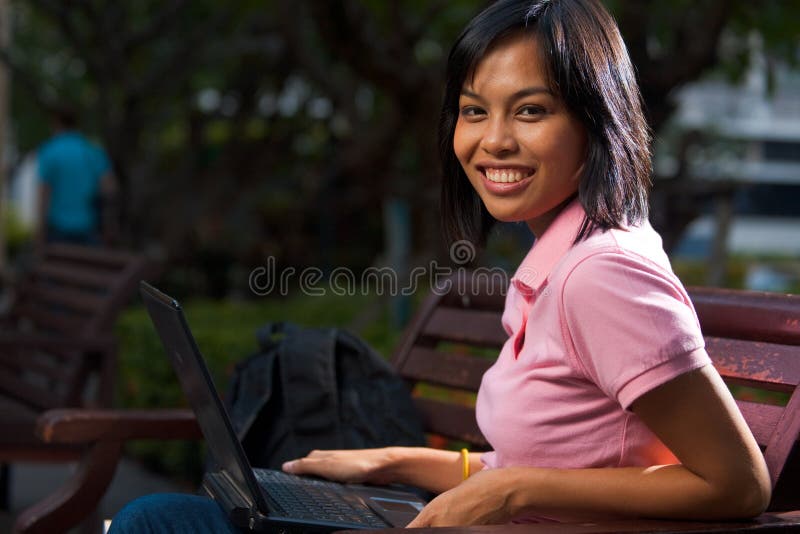 A cute college student smiling while using her laptop on a university campus bench. 20s female Asian Thai model of Chinese descent. A cute college student smiling while using her laptop on a university campus bench. 20s female Asian Thai model of Chinese descent.