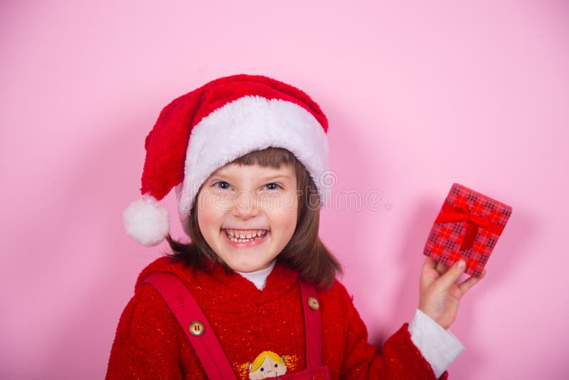 Cute smiling little girl in Santa hat and Christmas costume holding red gift box in studio on pink background. stock images Cute smiling little girl in Santa hat and Christmas costume holding red gift box in studio on pink background. stock images