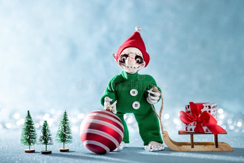 Cute Smiling Santas Helper Elf Holding Christmas Bauble and Pulling Christmas Gift on a Sledge. North Pole Christmas Scene.