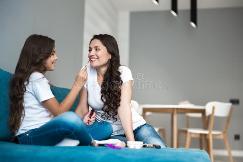 Cute teenager daughter applying lipstick to her young beautiful mother`s lips both smiling stock photo Cute teenager daughter applying lipstick to her young beautiful mother`s lips both smiling stock photo