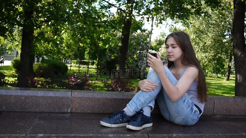 Cute teenager girl checking phone and waiting phone call sitting in park. Video footage HD shooting static camera.