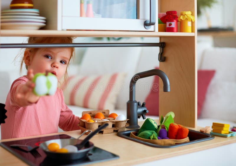 Cute toddler baby girl playing on toy kitchen at home, roasting eggs and treat you with apple slice, let`s share