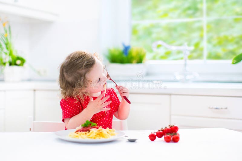 Cute toddler girl eating spaghetti in a white kitchen