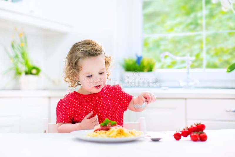 Cute toddler girl eating spaghetti in a white kitchen
