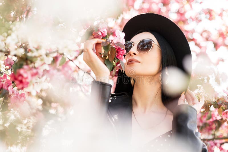 Cute woman in flowered park walks on nature. Lovely girl in elegant hat in trendy sunglasses in black fashionable outfit and pink
