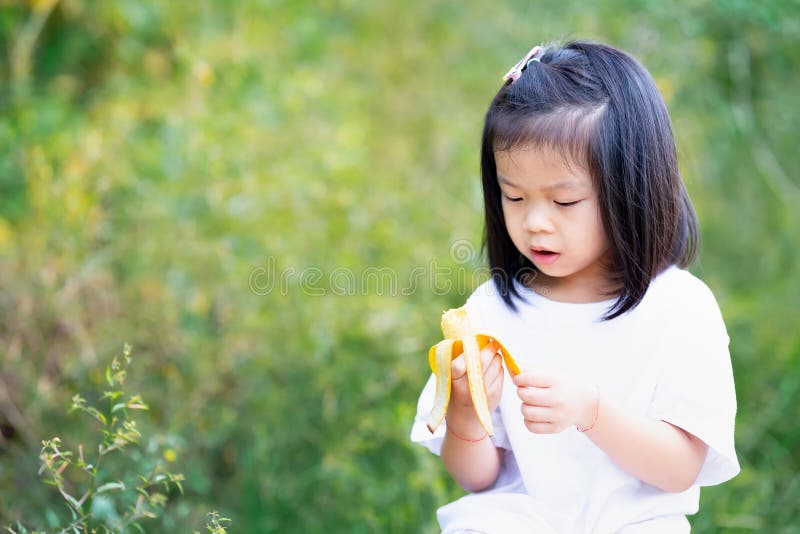 Cute 4 year old Asian girl is eating banana for breakfast. Child holds yellow fruit in her hand. Banana peeling kid.