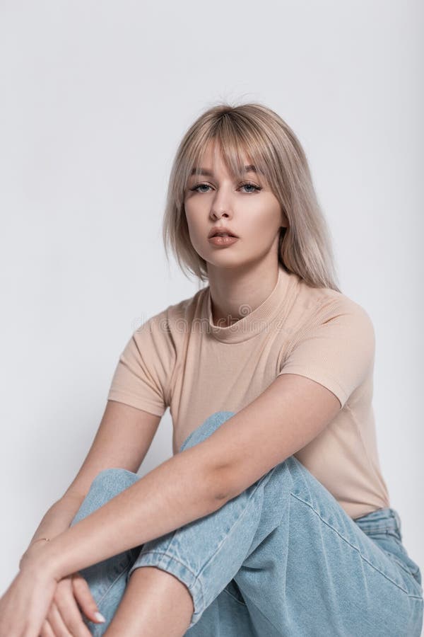 Cute young attractive woman model in fashionable youth clothes is sitting near a white vintage wall. Pretty glamorous blonde girl