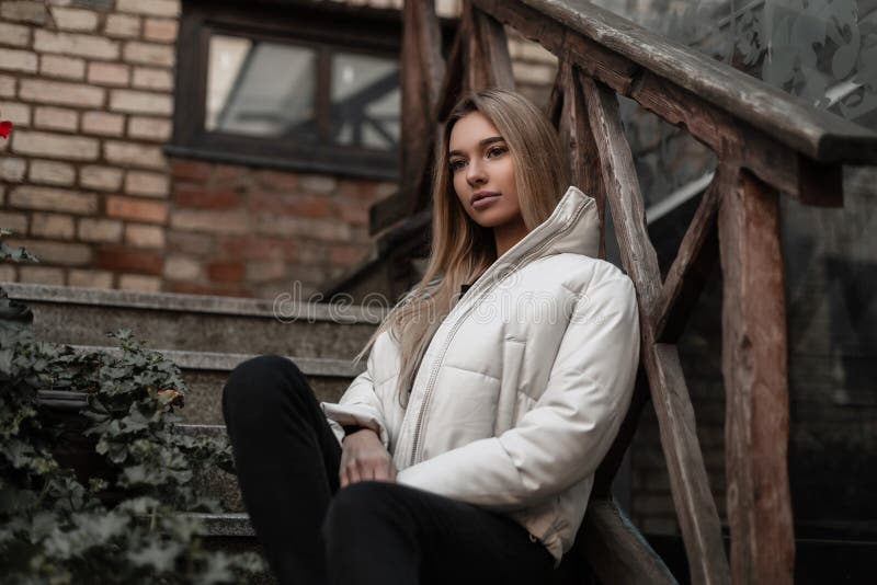 Cute young blond woman in a fashionable white jacket in stylish black jeans sits on a vintage stone staircase with wooden railing