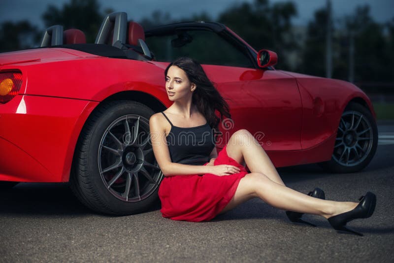 Cute young brunette woman sitting on the ground near the wheel of luxury red cabriolet car.