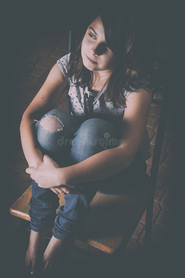 Dolce, triste bambina seduta con i piedi appoggiati su una sedia nel buio, la metà del suo viso nascosto dai capelli castani.