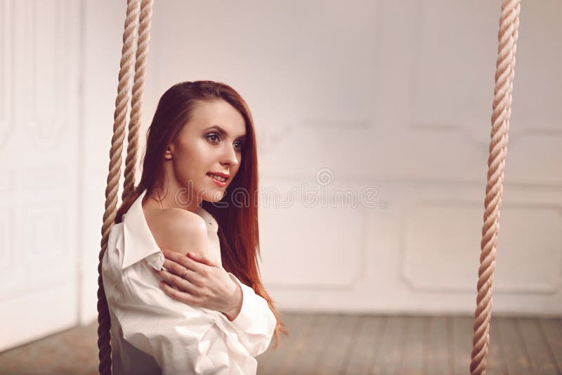 Cute young redhead woman with long hair sitting on swing in man`s shirt stock photography Cute young redhead woman with long hair sitting on swing in man`s shirt stock photography