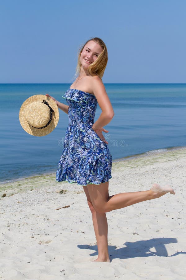 Cute young girl with a hat, in blue dress posing on the beach, sea background. Freedom and fun concept