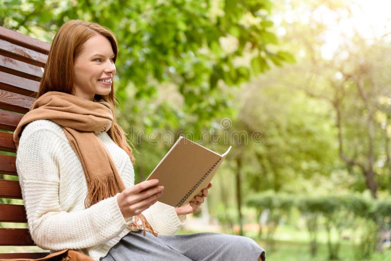 Joyful female student is studying in nature. She is sitting on bench and laughing. Girl is holding notebook and looking forward with interest. Joyful female student is studying in nature. She is sitting on bench and laughing. Girl is holding notebook and looking forward with interest