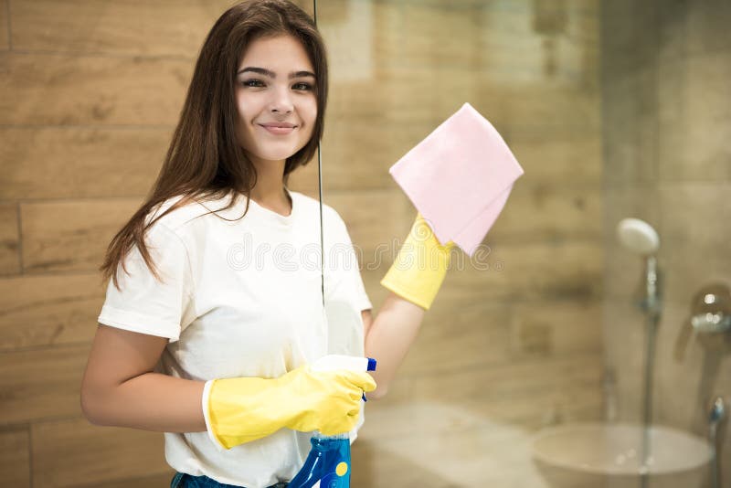 Cute young woman in yellow gloves spraying detergent spray while wiping dust off from the shower door with a rag in the
