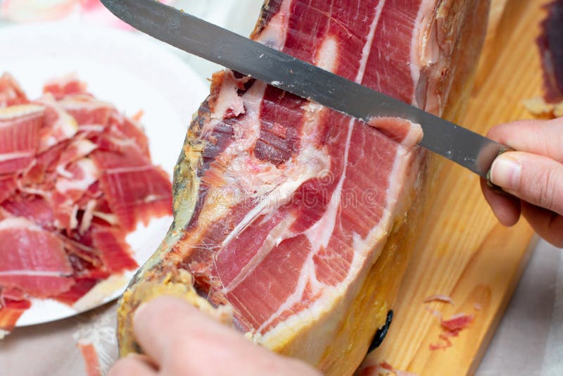 Cutting leg of Serrano ham with a knife, in the background a plate of split Serrano ham