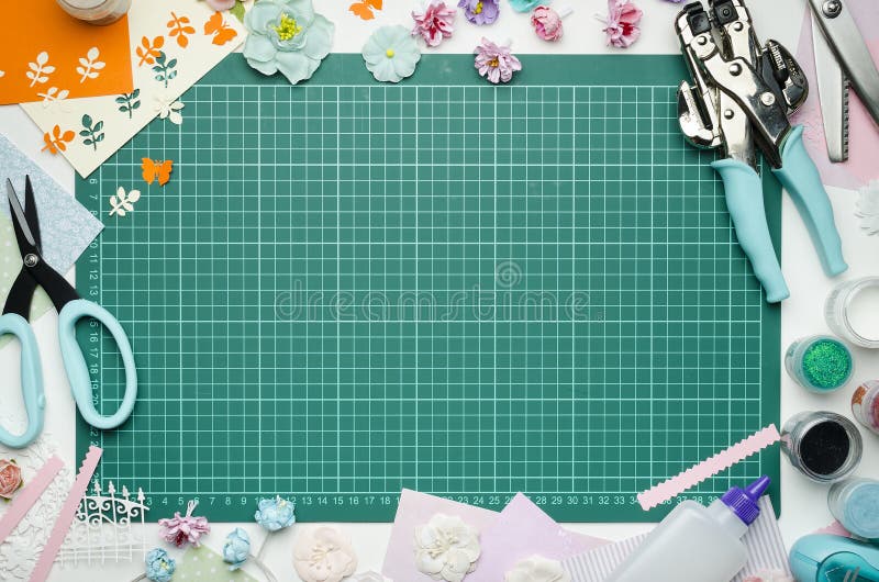 The cutting mat is surrounded by paper flowers, paper, tools and scrapbooking materials. Scrapbooking, top view, empty space in the center