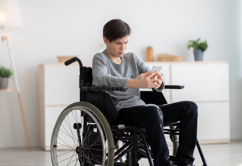 Cyberbullying. Stressed disabled teen boy in wheelchair looking at smartphone screen, being bullied online at home. Cyberbullying concept. Stressed disabled teen