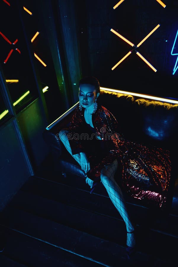 Cyberpunk shooting of model wearing red bathrobe with glitter sitting in leather sofa against neon