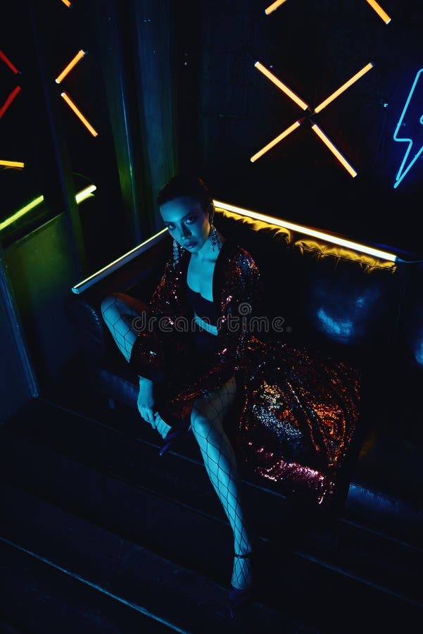 Cyberpunk shooting of model wearing red bathrobe with glitter sitting in leather sofa against neon