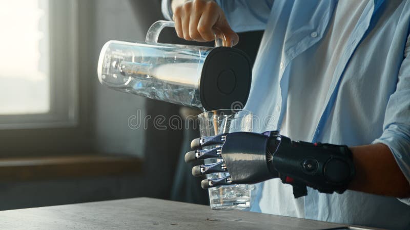 Cyborg τύπος με τεχνητό άκρο ρίχνει νερό στο ποτήρι από ένα μεγάλο μπολ και ποτά στέκεται στην κουζίνα στοκ εικόνα με δικαίωμα ελεύθερης χρήσης