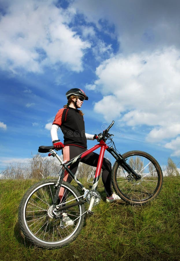 Female cyclist going up the slope with her bike. blue sky with clouds on the background