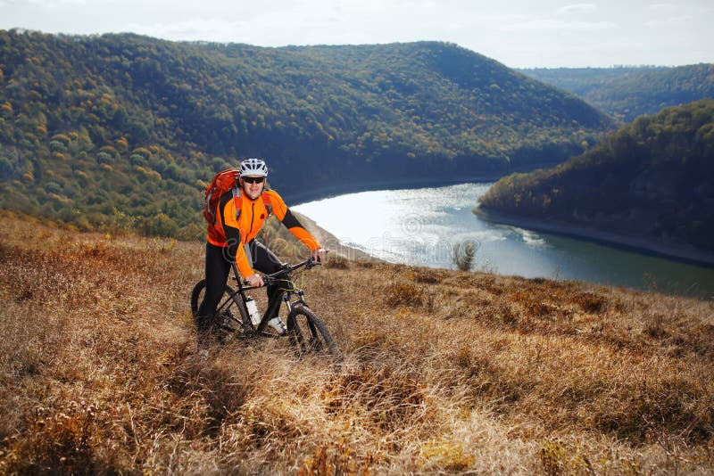 Cyclist in orange jacketr stands with his bike under river against beautiful landscape with mountain.
