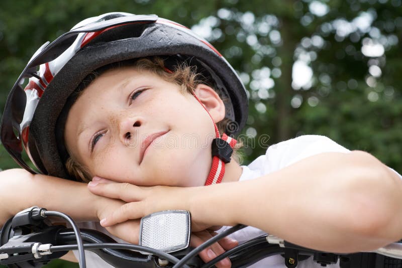 Cyklisten för den lilla kaukasiska pojken i skyddshjälmen satte huvudet på handledet på den cykel som ställer upp för kameran en arkivfoto