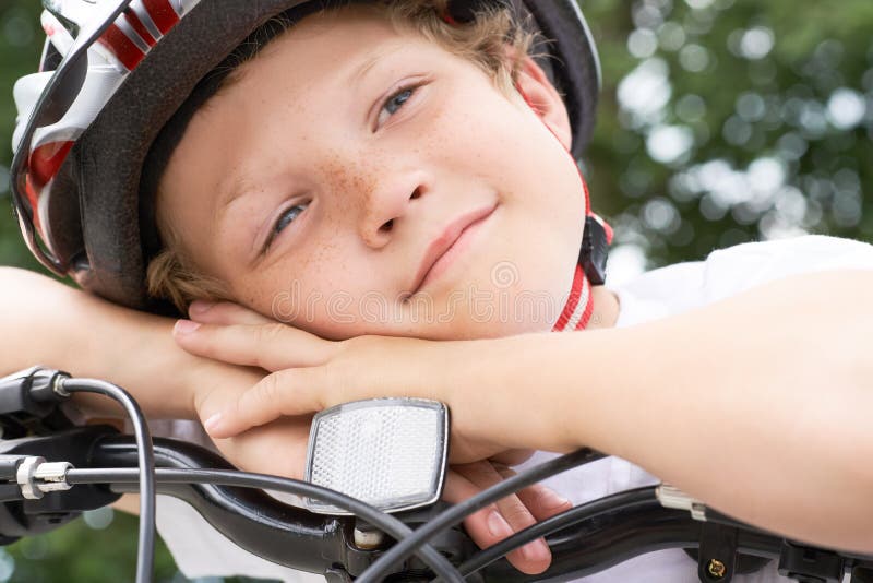 Cyklisten för den lilla kaukasiska pojken i skyddshjälmen satte huvudet på handledet på den cykel som ställer upp för kameran en royaltyfria foton
