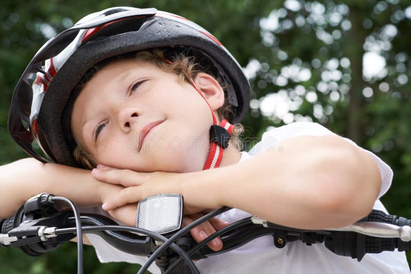 Cyklisten för den lilla kaukasiska pojken i skyddshjälmen satte huvudet på handledet på den cykel som ställer upp för kameran en arkivbilder