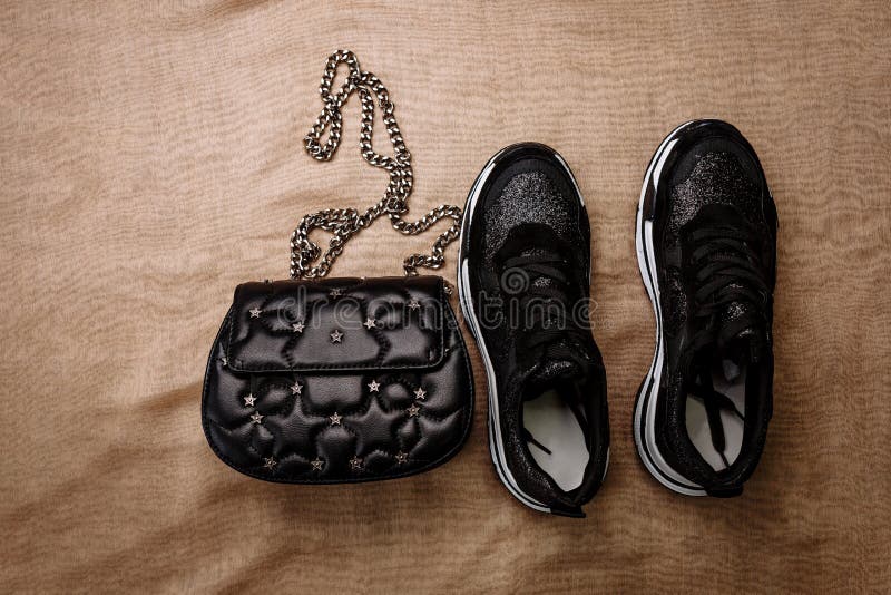 Czarni sneakers na białej gęstej podeszwie dekorującej z czernią błyskają i czarny sprzęgło z asteryskami na łańcuchu na beżu i fotografia stock Czarni sneakers na białej gęstej podeszwie dekorującej z czernią błyskają i czarny sprzęgło z asteryskami na łańcuchu na beżu i fotografia stock