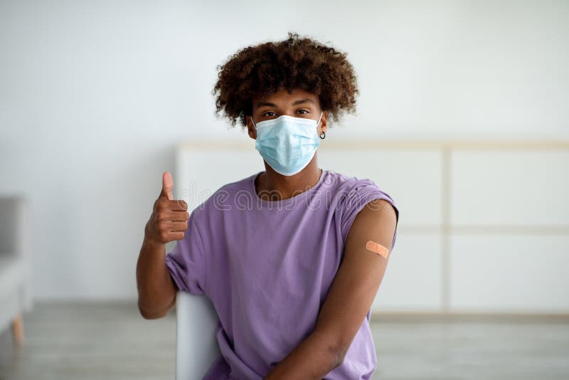 Czarny nastolatek maska na twarzy nosi plaster opatrunkowy na ramieniu po zastrzyku szczepionki, pokazując kciuk w domu