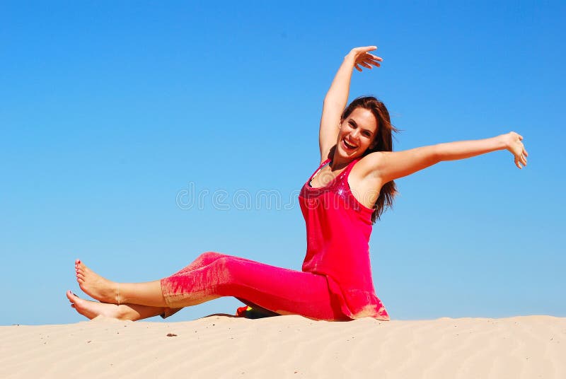 A beautiful young Caucasian woman in pink clothes with happy smiling facial expression sitting on a lonely sand dune in front of blue sky summer sky, raising up her arms, feeling free and great. A beautiful young Caucasian woman in pink clothes with happy smiling facial expression sitting on a lonely sand dune in front of blue sky summer sky, raising up her arms, feeling free and great.