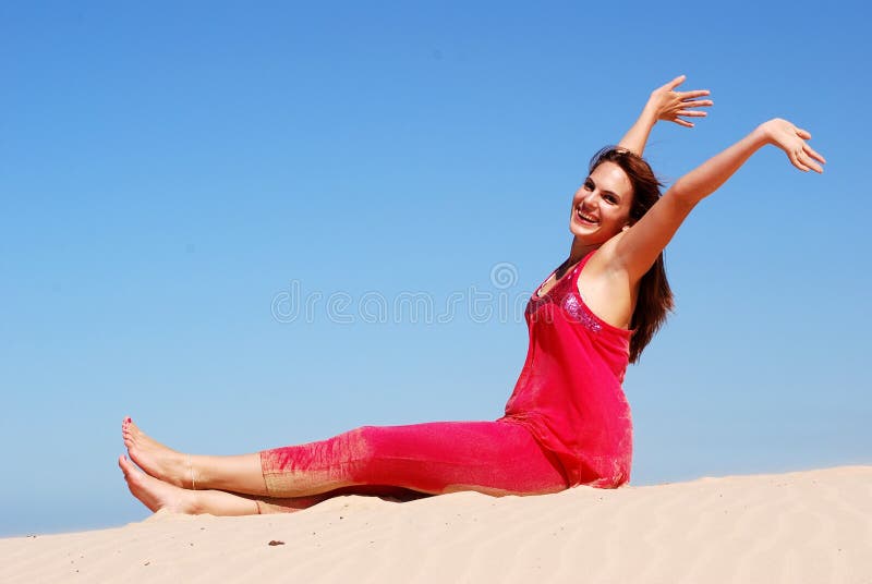 A beautiful young lady in pink clothes with happy smiling facial expression sitting on a lonely sand dune in front of blue sky summer sky, raising up her arms and feeling free. A beautiful young lady in pink clothes with happy smiling facial expression sitting on a lonely sand dune in front of blue sky summer sky, raising up her arms and feeling free.