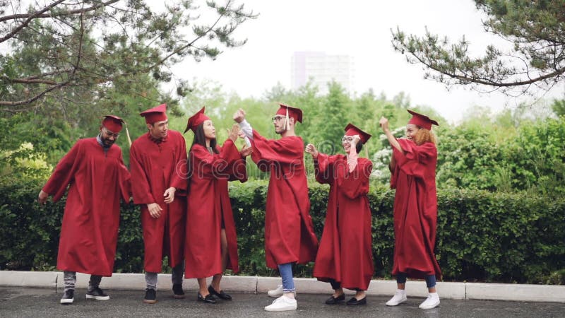 Cámara lenta del grupo multi-étnico de estudiantes en vestidos y de mortero-tableros que bailan y que ríen al aire libre en campu