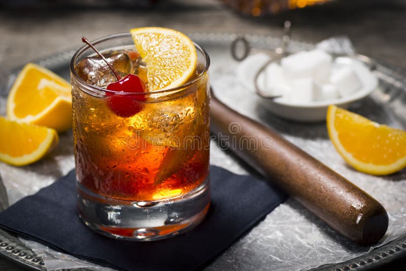 An Old Fashioned cocktail in a vintage bar setting with the ingredients and tools in the background. Old Fashioneds usually have some type of whiskey, bitters, muddled oranges, ice, and sometimes a cherry. An Old Fashioned cocktail in a vintage bar setting with the ingredients and tools in the background. Old Fashioneds usually have some type of whiskey, bitters, muddled oranges, ice, and sometimes a cherry.