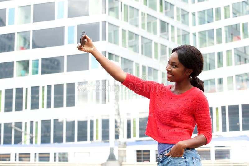 Côté de la jeune femme de couleur heureuse prenant la photo de selfie dans la ville image libre de droits Côté de la jeune femme de couleur heureuse prenant la photo de selfie dans la ville image libre de droits