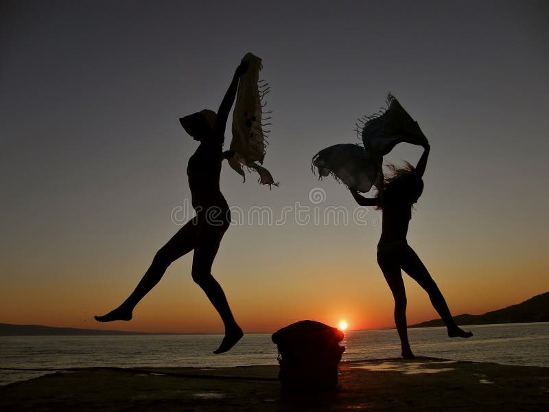 Silhouette of two women (mother and daughter - best friends) dancing - practice with a scarf by the Adriatic sea (mooring detail - cleat) in the beautiful orange sunset. Horizontal color photo. Serene beach atmosphere stock images, royalty-free photos and pictures