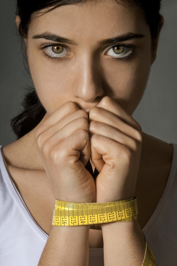 Moody photo of a beautiful anxious young girl trying to slim with diet. she is in danger because of forbidden eating disorder and hard dietary rules. Her hands are tied with a measuring tape. The woman looks sad, worried and concerned. Moody photo of a beautiful anxious young girl trying to slim with diet. she is in danger because of forbidden eating disorder and hard dietary rules. Her hands are tied with a measuring tape. The woman looks sad, worried and concerned.