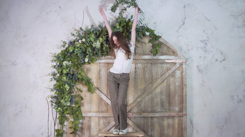 Dança de sorriso feliz da menina em uma escada portátil no fundo de uma porta de madeira bonita decorada com flores imagens de stock royalty free Dança de sorriso feliz da menina em uma escada portátil no fundo de uma porta de madeira bonita decorada com flores imagens de stock royalty free