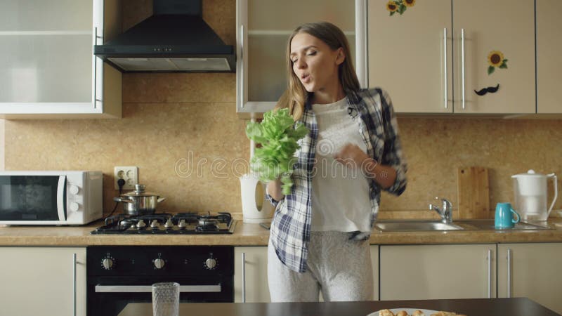 Dança engraçada nova alegre da mulher e canto com microfone da alface ao cozinhar o café da manhã