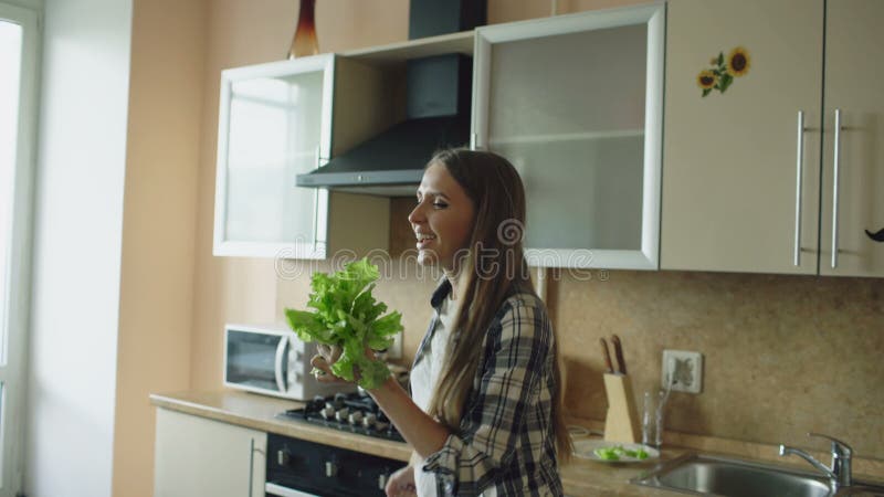 Dança engraçada nova alegre da mulher e canto com microfone da alface ao cozinhar o café da manhã