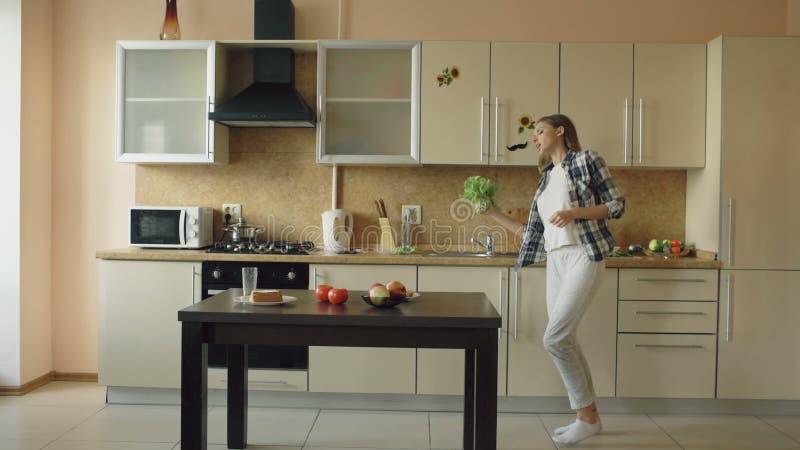 Dança engraçada nova alegre da mulher e canto com microfone da alface ao cozinhar o café da manhã