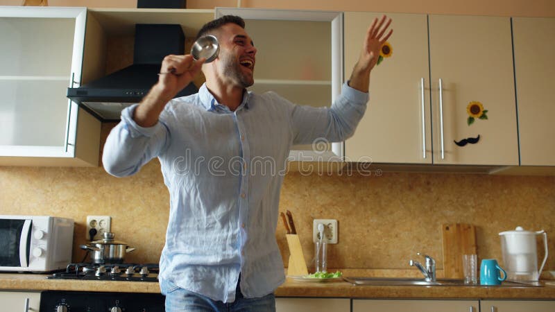 Dança engraçada nova atrativa do homem e canto com concha ao cozinhar na cozinha em casa fotografia de stock Dança engraçada nova atrativa do homem e canto com concha ao cozinhar na cozinha em casa fotografia de stock