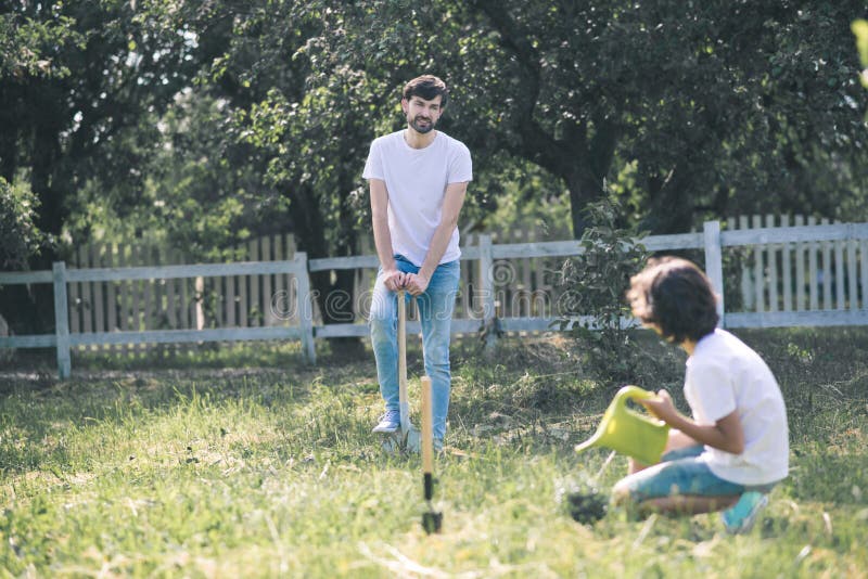 Dark-haired boy watering the plant, his father watching him stock photos