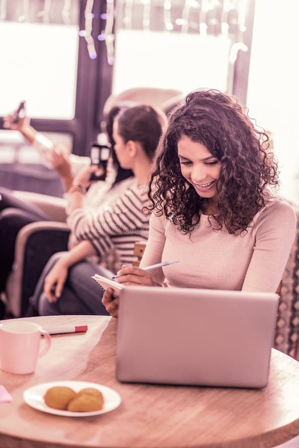 Dark-haired curly woman sitting with her laptop in bakery while eating tasty biscuits stock photos Dark-haired curly woman sitting with her laptop in bakery while eating tasty biscuits stock photos
