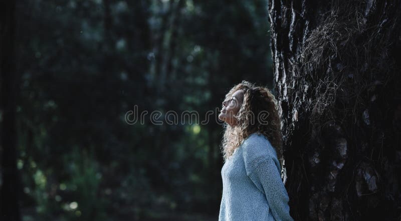 Dark image of woman standing against a tree trunk in oudoor nature leisure activity with woods in background. One female people
