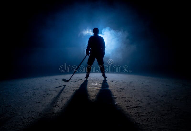 Dark silhouette of a male hockey player in a uniform, helmet and skates with a stick on the ice arena with smoke and