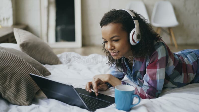 Das junge gelockte Afroamerikanerfrauengenießen hören Musik auf Laptop beim Lügen auf Bett