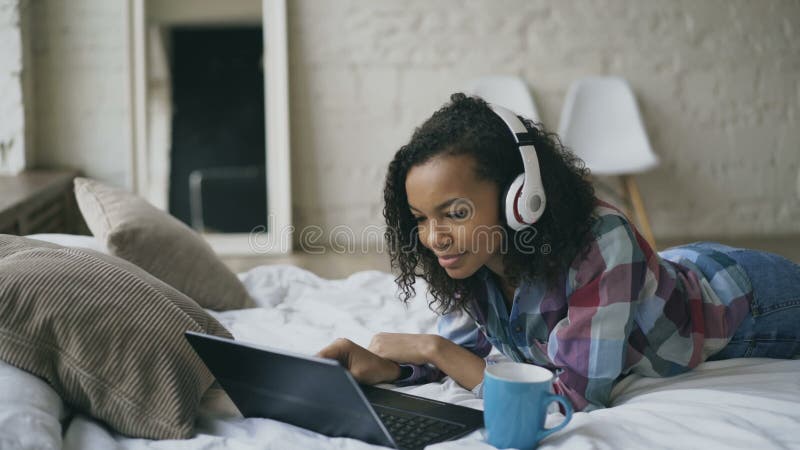 Das junge gelockte Afroamerikanerfrauengenießen hören Musik auf Laptop beim Lügen auf Bett